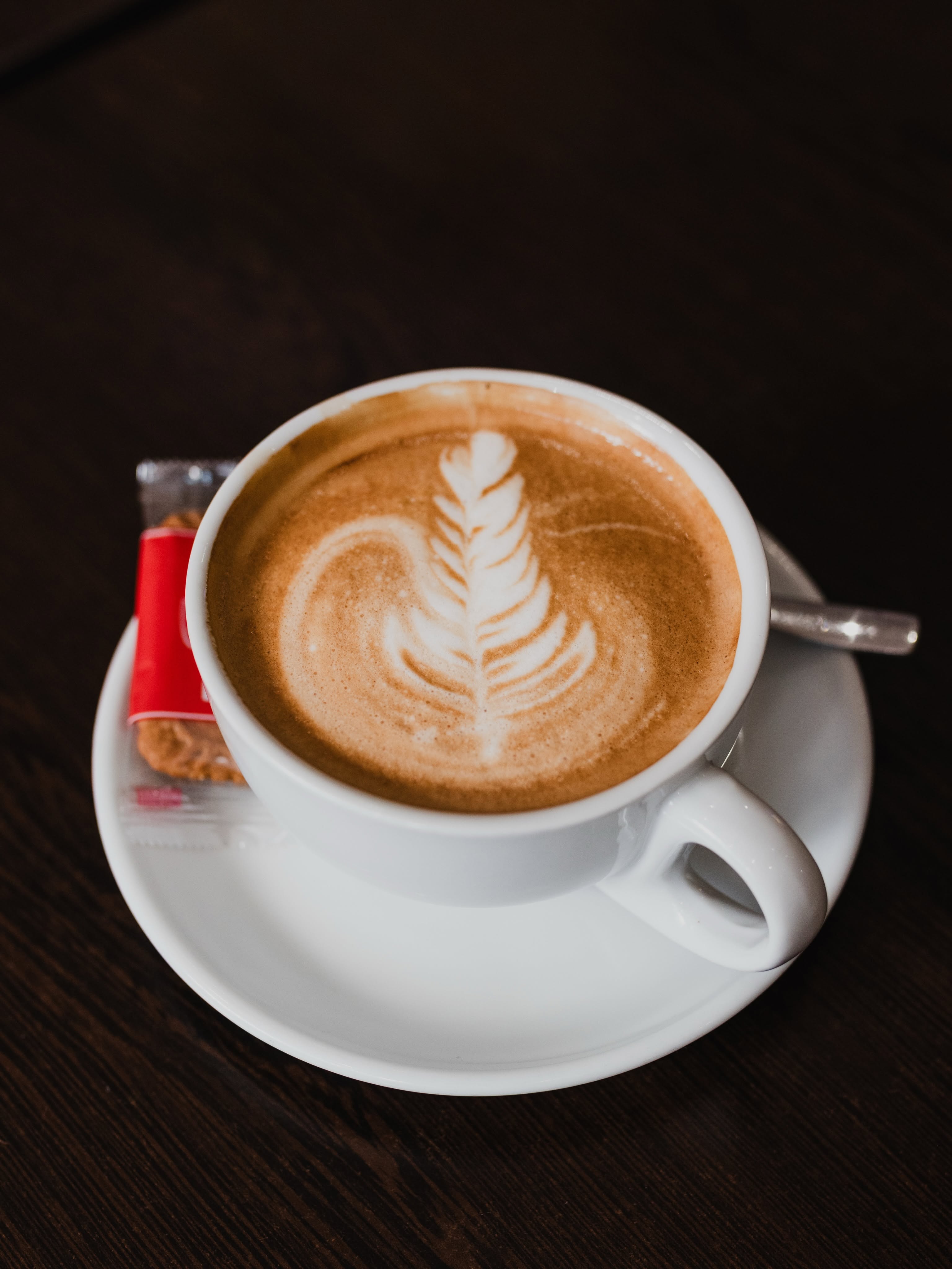 Aroma’s coffee in a complete white cup and saucer, with leaf-pattern foam, a spoon and biscuit